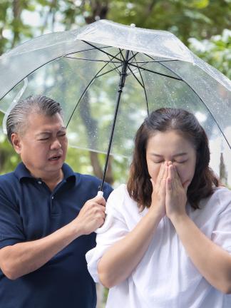 Image of an older adult woman blows her nose while her husband puts his hand around her. The both of them are under an umbrella