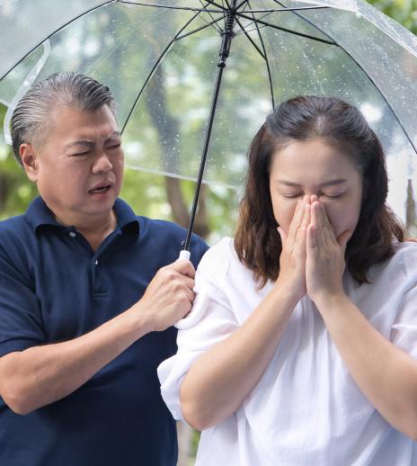 Photo of an older adult woman blows her nose while her husband puts his hand around her. The both of them are under an umbrella