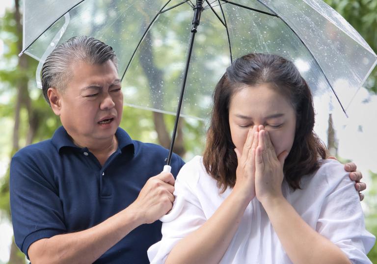 An older adult woman blows her nose while her husband puts his hand around her. The both of them are under an umbrella