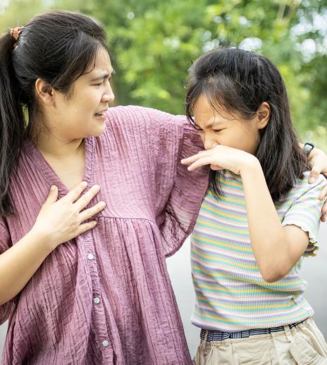 A mother has her hand around her daughter and looks distraught as her daughter looks disgustedly at her armpit, presumably because of body odor.