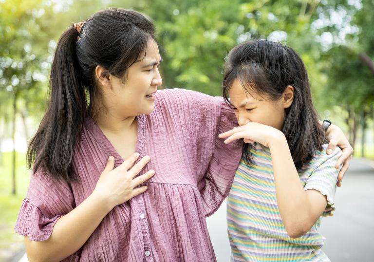 In a park, a mother has her hand around her daughter and looks distraught as her daughter looks disgustedly at her armpit, presumably because of body odor.