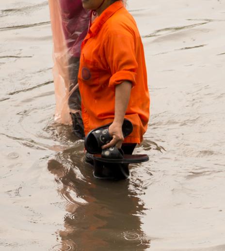 A person in an orange shirt wading through floodwater while holding her slippers in one hand.
