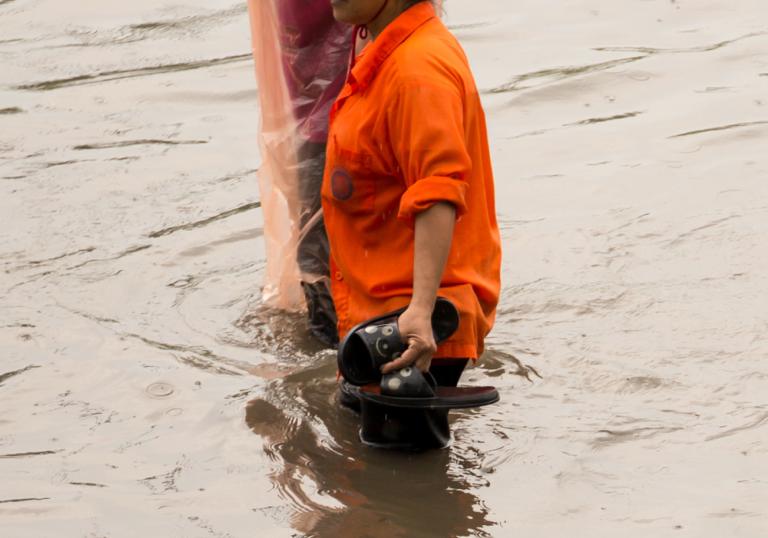 A person in an orange shirt wading through floodwater while holding their slippers in one hand.